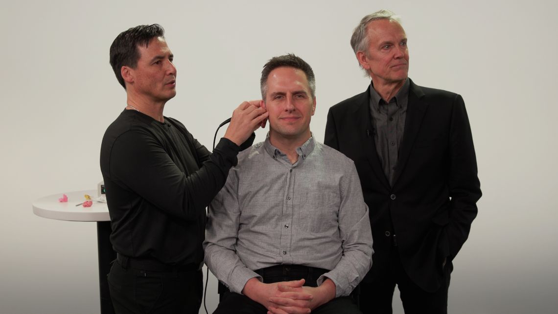 Two men watching a screen as he uses a video otoscope in seated man's ear.