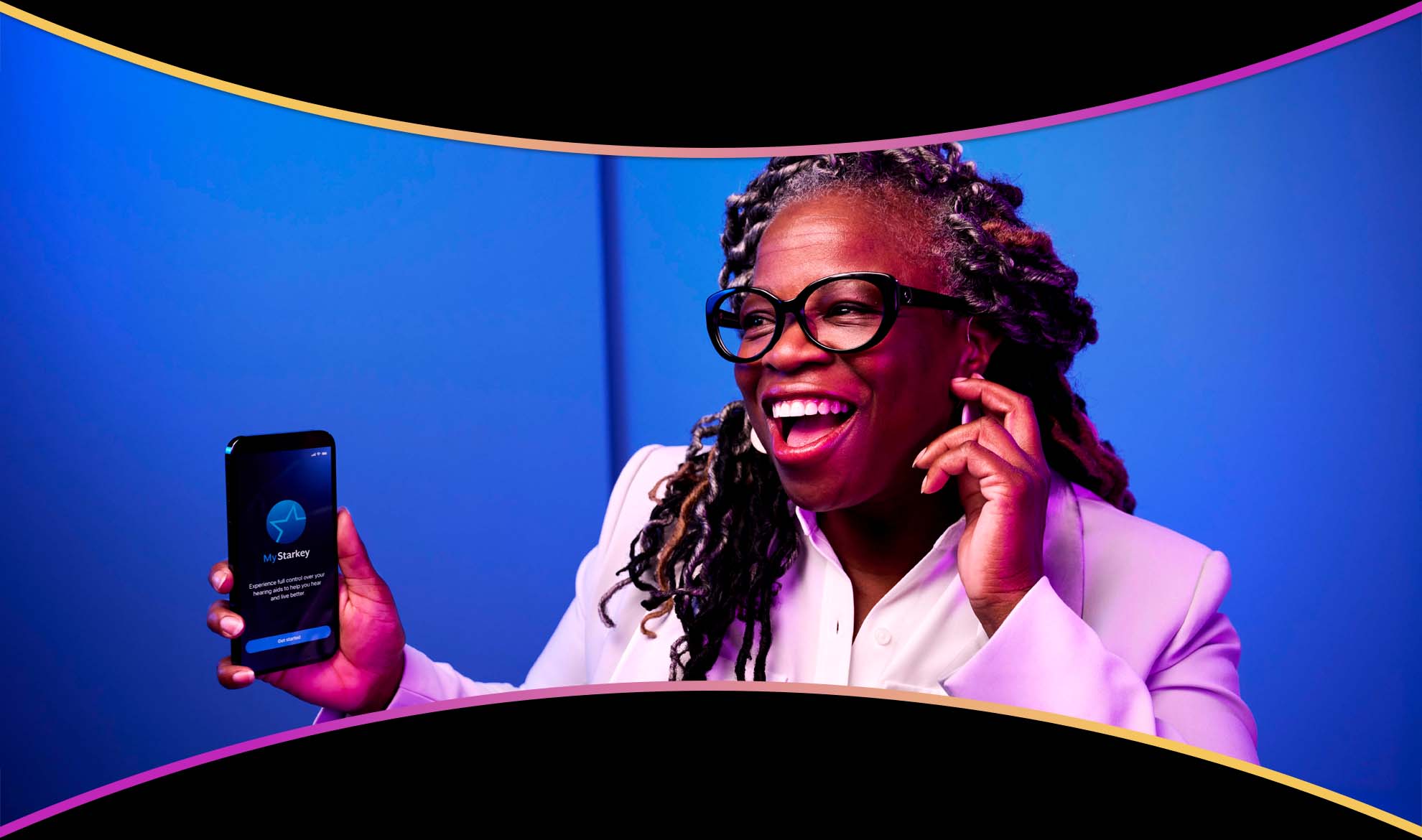 A smiling woman wearing hearing aids holding up her phone showing the My Starkey app