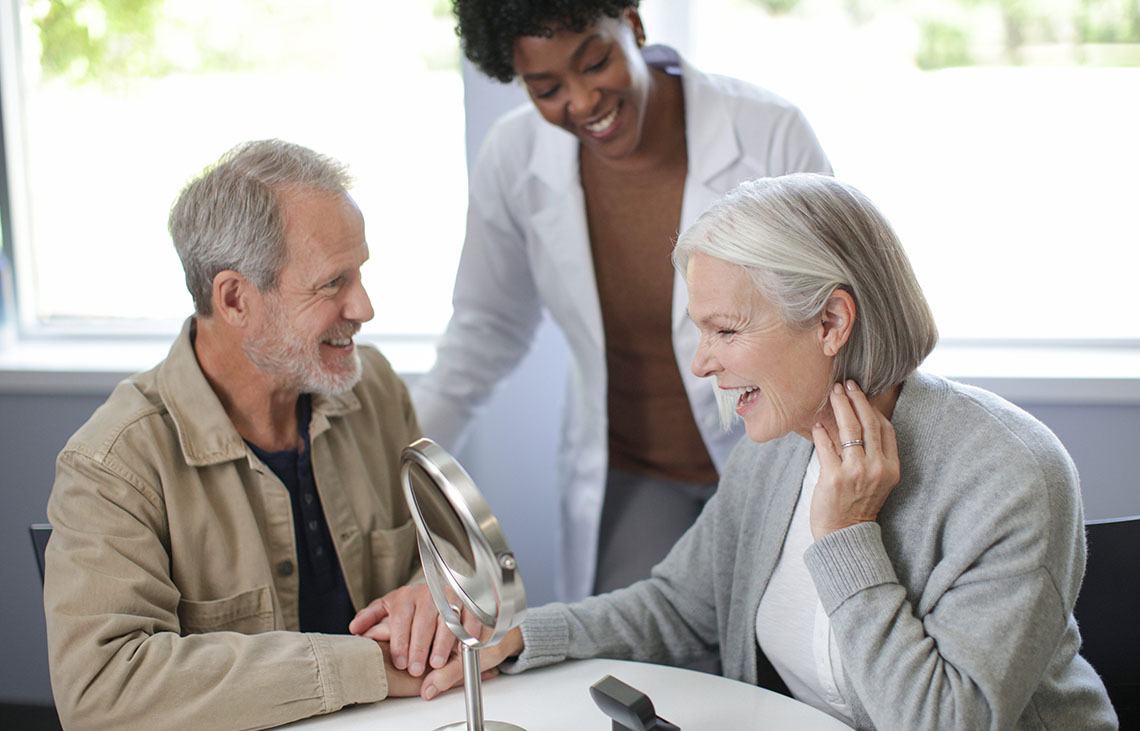 A hearing professional and a man watching a woman trying on a hearing aid