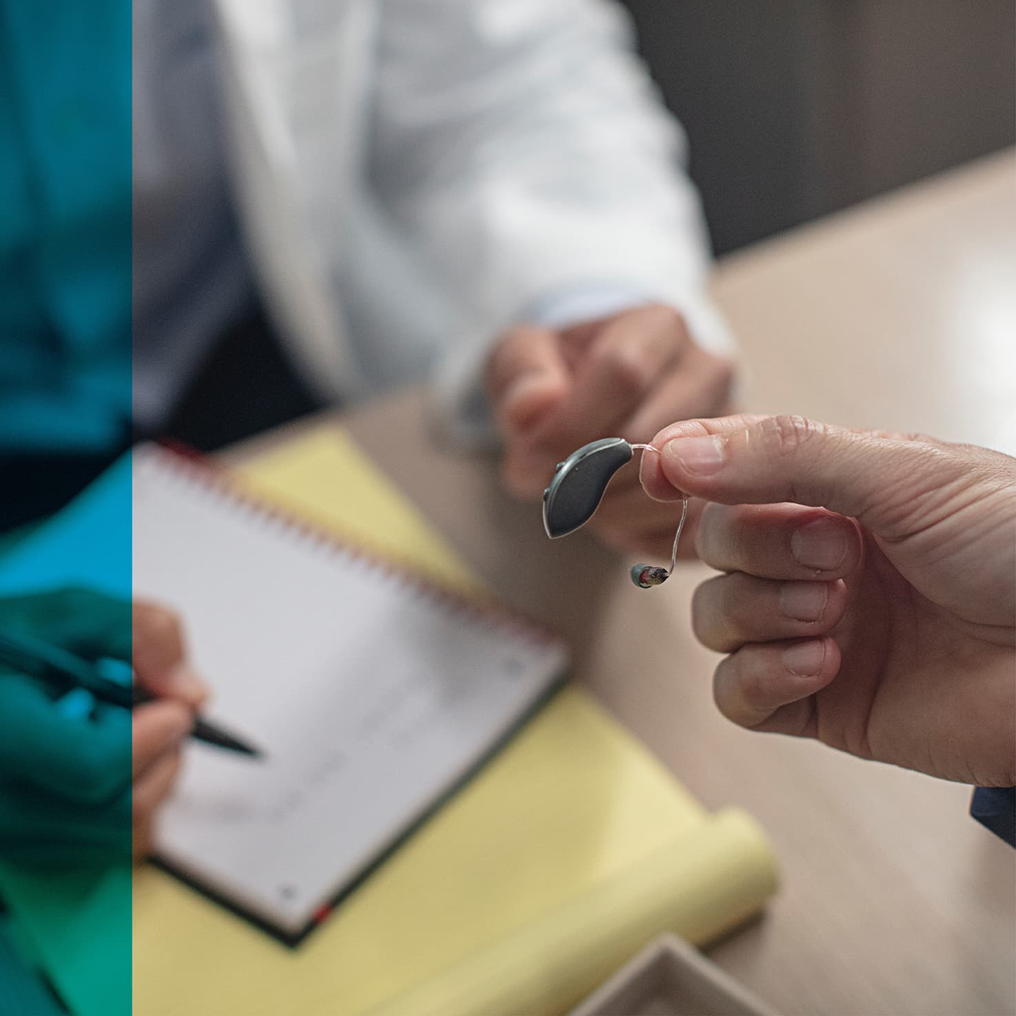 person's hand holding a hearing aid across the desk from an audiologist who takes notes